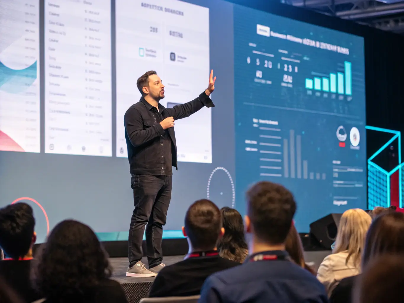 A photograph of a speaker presenting on stage at a technology conference in Manchester, UK, with a large screen displaying data visualizations.
